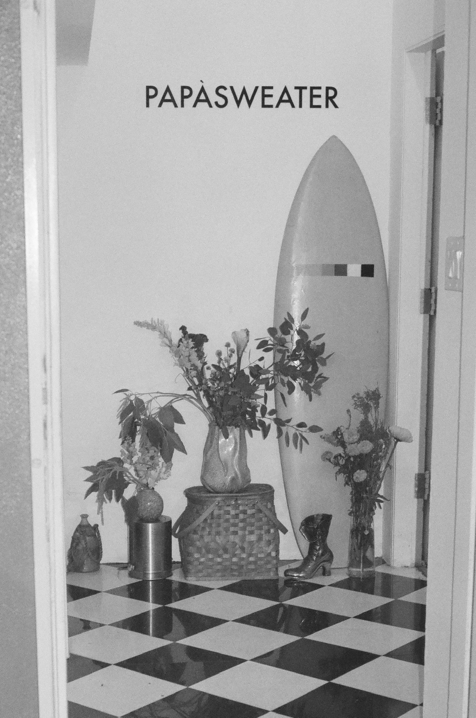 Inside Papasweater studio: flowers, a surfboard, and a vintage boot on a black-and-white checkerboard floor.
